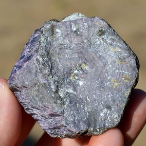 May include: A raw, hexagonal-shaped mineral specimen featuring a deep purple and lavender color palette. The surface displays a complex, crystalline texture with visible striations, metallic silver inclusions, and small patches of earthy tan mineral deposits.