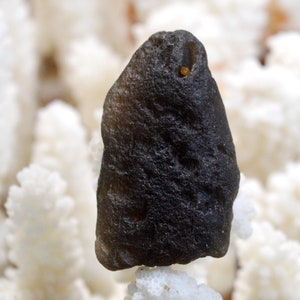 May include: A dark gray, irregularly shaped stone with a small, light brown spot on the surface. The stone is resting on a white, textured surface.