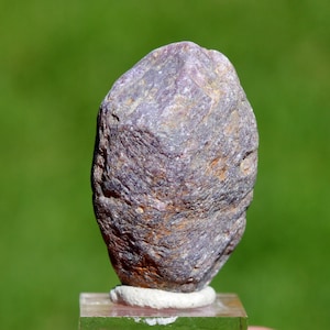 Könnte beinhalten: Ein rauer, unregelmäßig geformter Stein mit einer Mischung aus lila, braunen und grauen Farbtönen. Der Stein ist auf einem transparenten, rechteckigen Sockel mit einer weißen, kreisförmigen Halterung montiert. Der Hintergrund ist ein verschwommenes Grün.