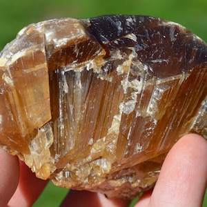 May include: A raw, natural mineral specimen with a crystalline structure. The stone displays a range of warm colors, from light amber to deep brown, with areas of white and translucent sections. The surface has a rough, textured appearance.