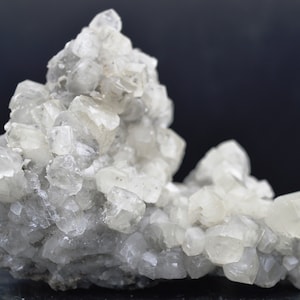 Puede incluir: Un grupo de cristales de calcita claros y blancos sobre una matriz gris. Los cristales son afilados y bien formados, con un brillo v&iacute;treo.