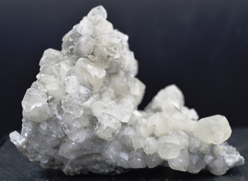 Puede incluir: Un grupo de cristales de calcita claros y blancos sobre una matriz gris. Los cristales son afilados y bien formados, con un brillo v&iacute;treo.