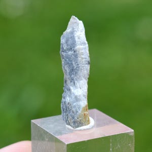 Op de afbeelding: Een ruw, lichtgrijs mineraal specimen, verticaal georiënteerd, gemonteerd op een heldere, vierkante basis. Het mineraal heeft een ruw, getextureerd oppervlak met hints van beige en donkerder grijs. De achtergrond is een vage groene kleur.