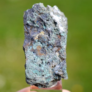 May include: A rough, gray, and green rock with blue and white mineral inclusions. The rock is on a clear stand.
