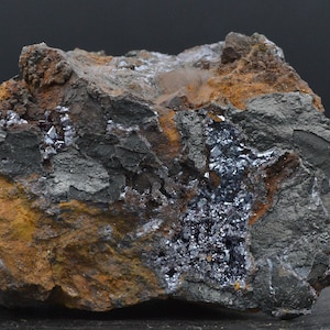 May include: A close-up of a rough, textured rock formation. The rock displays a mix of dark gray, rusty orange, and black colors. Crystalline structures are visible within the rock's crevices, adding a sparkling contrast to the earthy tones.