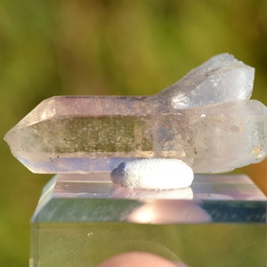 May include: A clear quartz crystal with a double termination, showing a light pink hue. The crystal is sitting on a small, white, round stand.