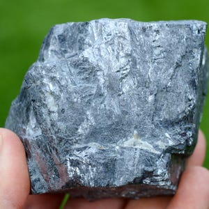 May include: A hand holds a rough, gray rock with a metallic sheen. The rock's surface is textured with visible lines and variations in color, ranging from dark gray to silver. The background is a blurred green.