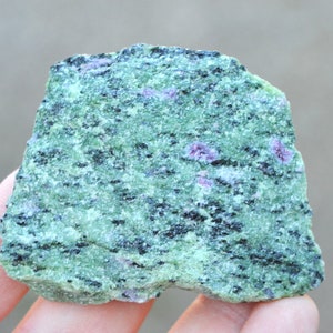 Op de afbeelding: Een ruwe, groene en zwarte steen met kleine, roze insluitsels. De steen heeft een gemarmerd, getextureerd oppervlak.