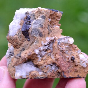 Puede incluir: Un espécimen mineral natural y rugoso que presenta una mezcla de colores y texturas. La roca muestra una matriz de tonos marrones y beige, con parches de cristales blancos y grises. Una formación cristalina oscura está incrustada en la piedra.