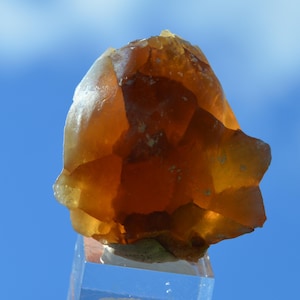 May include: A translucent, amber-colored mineral specimen with a rough, faceted surface. The stone is displayed against a bright blue sky, showcasing its natural, earthy tones and crystalline structure. The mineral appears to be a type of quartz or similar gemstone.