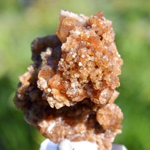 Könnte beinhalten: Nahaufnahme eines Clusters aus orange-braunen Kristallen. Die Kristalle haben eine raue, strukturierte Oberfläche und sind in einer natürlichen Formation zusammengefasst. Der Hintergrund ist verschwommen, mit grünen und weißen Farbtönen.
