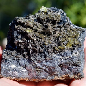 Könnte beinhalten: Ein dunkelgrauer Stein mit eingebetteten grünen und klaren Kristallen. Der Stein ist etwa handgroß.