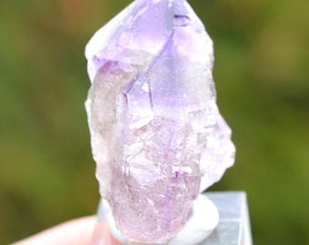 May include: A purple amethyst crystal point with a clear, glassy appearance. The crystal is on a small, clear display stand.