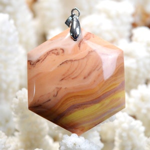 May include: A hexagonal pendant with a polished, swirled pattern in shades of peach, brown, and yellow. The gemstone pendant is suspended from a silver-colored bail. The background is a blurred white.