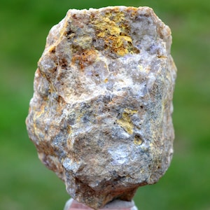 May include: A rough, irregularly shaped rock with a mix of colors. The stone features a base of light gray and tan, with patches of yellow and brown. The surface has a textured appearance, with visible mineral deposits. The rock is likely a natural geological specimen.