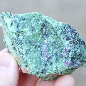 Può includere: Una roccia ruvida, verde e nera con piccole macchie viola. La roccia ha un aspetto screziato ed è un esemplare naturale.