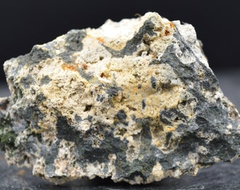 May include: A close-up of a rough, textured rock sample. The rock has a mix of colors, including gray, beige, and black, with small orange and green accents. The surface is uneven and porous, suggesting a natural mineral formation.