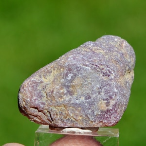 Könnte beinhalten: Nahaufnahme eines rauen, natürlichen Edelsteins mit einer Mischung aus lila, braunen und beigen Farbtönen. Der Stein hat eine strukturierte Oberfläche und wird auf einem transparenten, rechteckigen Ständer vor einem verschwommenen grünen Hintergrund präsentiert.