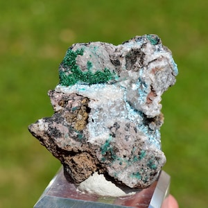 Könnte beinhalten: Ein rauer, grauer Stein mit grünen und blauen Kristallen. Der Stein befindet sich auf einem durchsichtigen Ausstellungsständer.
