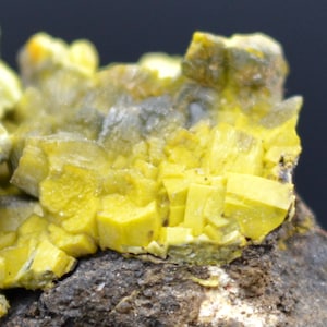 Op de afbeelding: Close-up van een geel mineraalmonster met een ruwe, kristallijne textuur. Het mineraal is geclusterd in een dichte formatie, met enkele zichtbare individuele kristallen. Het monster is geplaatst tegen een donkere achtergrond.