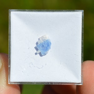 May include: A small, light blue crystal with white striations, displayed on a white background in a clear plastic square.