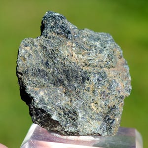 May include: A dark green, rough, and textured rock specimen. The rock has a slightly metallic sheen and appears to be a type of igneous rock.