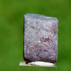 Könnte beinhalten: Ein rechteckiger, lila-grauer Edelstein mit rauer, strukturierter Oberfläche. Der Stein ist vor einem verschwommenen grünen Hintergrund platziert. Der Edelstein ruht auf einem kleinen, transparenten Ständer.