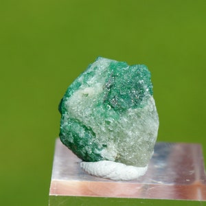 May include: A rough, green emerald crystal with a white, cloudy matrix. The crystal is on a white display stand.