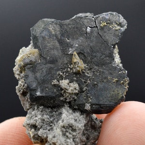 Op de afbeelding: Een donkergrijs mineraalmonster met een metallic glans. Het monster heeft een scherpe, hoekige vorm en is ingebed in een matrix van lichter grijs gesteente. Er zijn kleine, goudkleurige kristallen verspreid over het hele monster.