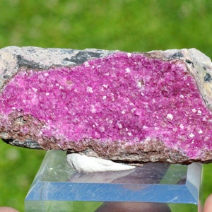 May include: A pink cobalt mineral specimen with a rough, natural surface. The mineral is embedded in a grey rock matrix.