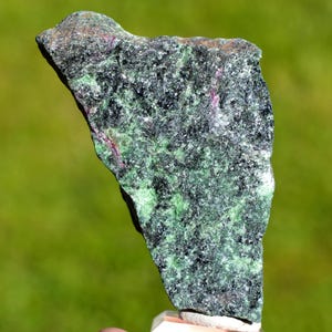 May include: A rough-cut stone with a dark green and black speckled pattern, accented with small red veins. The stone has a natural, uneven shape and texture, with a light-coloured base.