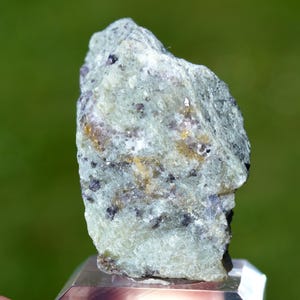 May include: A raw mineral specimen featuring a pale green, crystalline base with embedded dark purple and golden-yellow inclusions. The stone has a rough, jagged texture with visible facets and a translucent quality throughout its irregular structure.