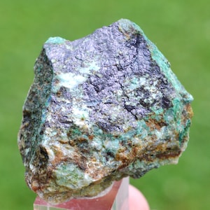 May include: A close-up of a rough mineral specimen. The rock has a dark, metallic sheen with areas of teal, green, and brown. The mineral is resting on a clear, rectangular base, with a blurred green background.