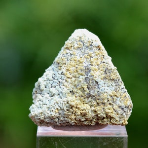 May include: A rough, triangular shaped rock with a white and yellow colour pattern. The rock is sitting on a clear glass display stand.