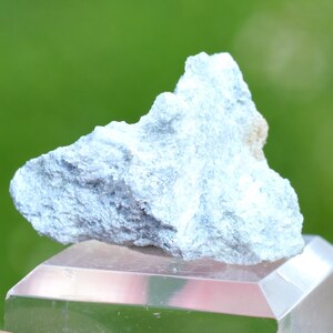 May include: A light blue mineral specimen with a rough, textured surface. The mineral sits atop a clear, geometric display stand, with a blurred green background.