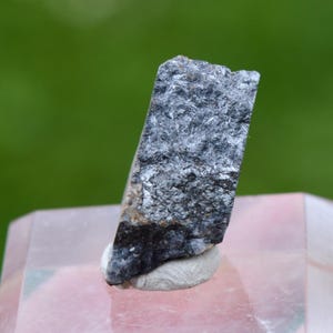 Op de afbeelding: Een close-up van een mineraalmonster. De steen heeft een rechthoekige vorm met een ruw, getextureerd oppervlak. Het bovenste gedeelte is lichtgrijs van kleur en gaat over in donkerder grijs en zwart aan de basis. Het exemplaar is gemonteerd op een kleine, lichtgekleurde basis.