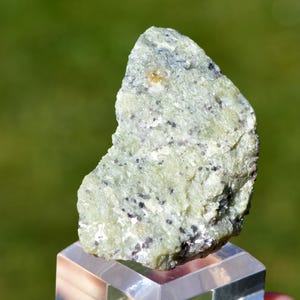 May include: A raw, pale green mineral specimen with a rough, crystalline texture and scattered dark grey to black speckles throughout its surface. The stone features a jagged, irregular shape with visible light-reflecting facets and mineral inclusions.