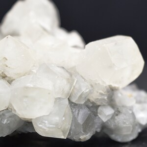 Puede incluir: Un grupo de cristales de calcita blancos con una superficie rugosa y gris. Los cristales son transl&uacute;cidos y tienen un ligero brillo iridiscente.