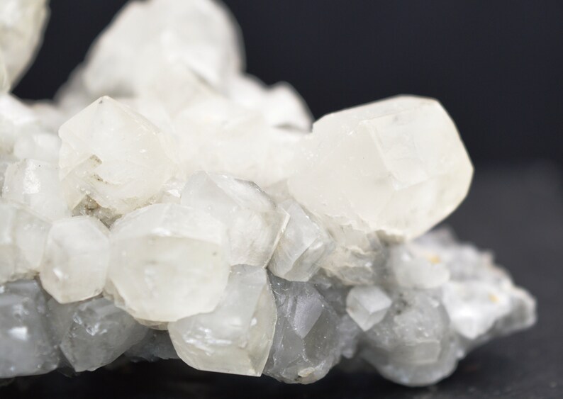 Puede incluir: Un grupo de cristales de calcita blancos con una superficie rugosa y gris. Los cristales son transl&uacute;cidos y tienen un ligero brillo iridiscente.