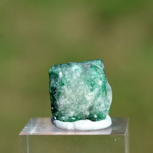 May include: A green emerald gemstone with a rough, natural surface. The emerald is displayed on a white pedestal.