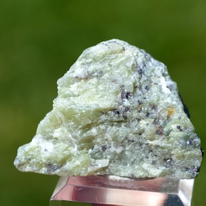 May include: A rough, triangular-shaped mineral specimen featuring a pale green, crystalline texture with scattered dark grey and black inclusions throughout the surface. The stone displays a layered, fibrous appearance and rests on a clear base.