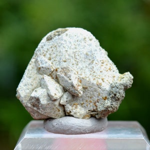 May include: A white, crystalline mineral specimen with a rough, textured surface. The mineral is displayed on a small, grey pedestal.
