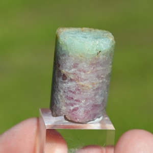 Puede incluir: Un espécimen de cristal verde y rosa con una apariencia en capas. El cristal se muestra en un soporte transparente.