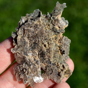 May include: A mineral specimen held in a hand, with a layered, crystalline structure. The colours are grey, brown, and yellow. The edges are jagged and irregular, with a rough texture. The background is a blurred green. The mineral is likely a type of mica.