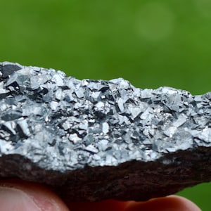 Può includere: Primo piano di un campione minerale grigio scuro con una struttura cristallina. La superficie è ricoperta da piccoli cristalli riflettenti dall'aspetto metallico. Lo sfondo è verde sfocato, che suggerisce un ambiente esterno.