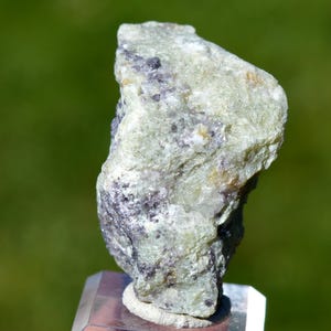 May include: A raw, irregular mineral specimen featuring a pale, translucent mint-green base color with scattered inclusions of deep purple and white crystalline clusters throughout its textured, jagged surface, mounted on a clear acrylic base.