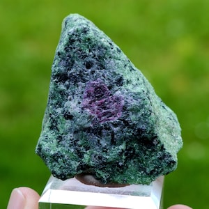 May include: A green and purple rough gemstone with a rough, irregular shape. The gemstone is on a clear display stand.