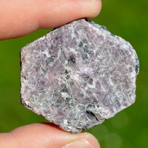 Puede incluir: Una piedra preciosa hexagonal rugosa de color gris púrpura con inclusiones negras. La piedra preciosa está siendo sostenida por una mano.