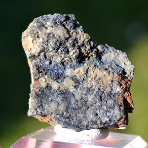 Può includere: Primo piano di una roccia grigio scuro con piccoli cristalli di quarzo trasparenti incastonati. La roccia ha una superficie ruvida e testurizzata ed è ricoperta da uno strato di polvere fine grigia.