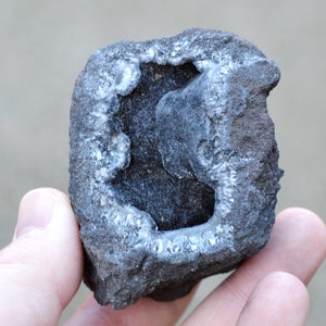 May include: A dark gray rock with a hollow center lined with white crystals. The rock has a rough, textured surface.